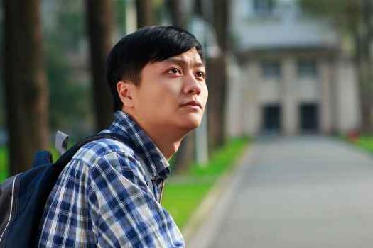 Asian man with backpack and plaid shirt looking up.